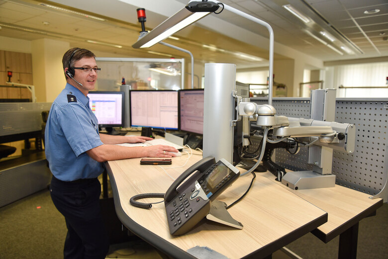 Hier gehen die Notrufe über die 110 ein: Ein Polizist in Uniform steht an einem Stehschreibtisch mit mehreren Monitoren. Er trägt ein Headset.