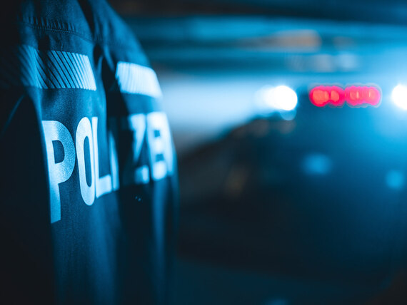 Der Schriftzug »Polizei« steht auf dem Rücken der Jacke eines Einsatzbeamten, im Hintergrund ist das Blaulicht eines Streifenwagens zu sehen.