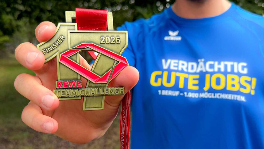 Ein Mitarbeiter der Polizei Sachsen zeigt die Finisher-Medaille der 17. REWE Teamchallenge 2026. Der Mitarbeiter trägt ein blaues Laufshirt mit der Karriere-Kampagne »Verdächtig gute Jobs! 1 Beruf – 1.000 Möglichkeiten« der sächsischen Polizei.